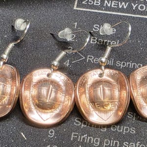 May include: Four copper penny earrings with silver-colored hooks. Each earring features a penny with the Lincoln Memorial design. The earrings are displayed on a dark, textured surface.