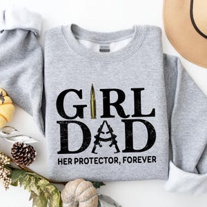 Pode incluir: Um moletom cinza claro com as palavras "GIRL DAD" em preto, com uma bala dourada substituindo o "I" em "GIRL" e a letra "A" em "DAD" formada por dois rifles. O texto "HER PROTECTOR, FOREVER" est&aacute; abaixo.