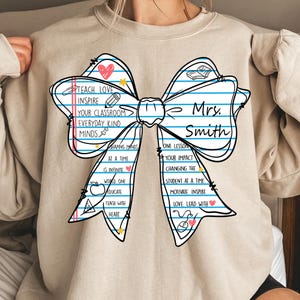 May include: Beige sweatshirt featuring a large bow design with the text "Teach Love Inspire" and "Mrs. Smith". The bow includes school-themed graphics and inspirational phrases. A perfect gift for teachers.