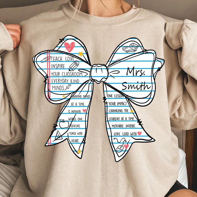 May include: Beige sweatshirt featuring a large bow design with the text "Teach Love Inspire" and "Mrs. Smith". The bow includes school-themed graphics and inspirational phrases. A perfect gift for teachers.