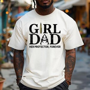 Pode incluir: Camiseta creme com o texto "GIRL DAD" em letras grandes, pretas e em negrito. A palavra "DAD" apresenta um design de dois rifles cruzados. O texto "HER PROTECTOR, FOREVER" est&aacute; abaixo.
