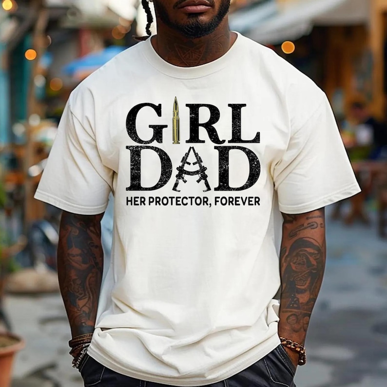 Pode incluir: Camiseta creme com o texto "GIRL DAD" em letras grandes, pretas e em negrito. A palavra "DAD" apresenta um design de dois rifles cruzados. O texto "HER PROTECTOR, FOREVER" est&aacute; abaixo.