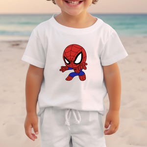 May include: White t-shirt featuring a cartoon Spider-Man graphic. The Spider-Man character is depicted in red, blue, and black, with large white eyes. The shirt is short-sleeved and appears to be made of a soft material.