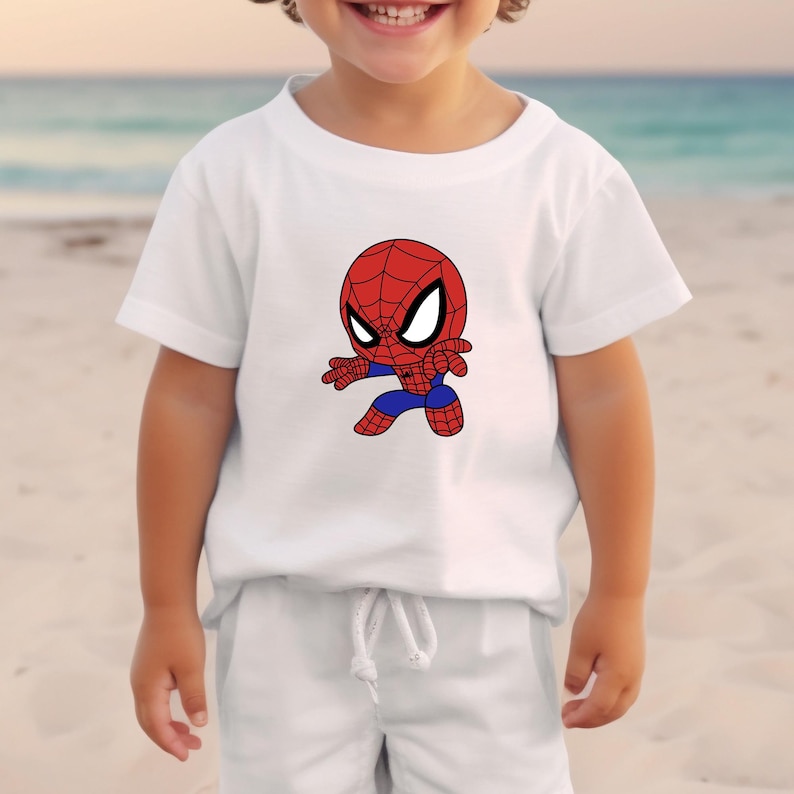 May include: White t-shirt featuring a cartoon Spider-Man graphic. The Spider-Man character is depicted in red, blue, and black, with large white eyes. The shirt is short-sleeved and appears to be made of a soft material.