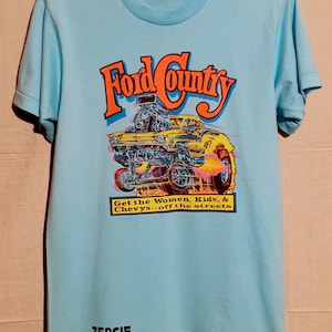 May include: Pale blue t-shirt featuring a yellow hot rod car graphic with the text "Ford County" in orange and blue. The shirt also has the text "Get the Women, Kids, & Chevys.. off the streets" and "ZERCIE".
