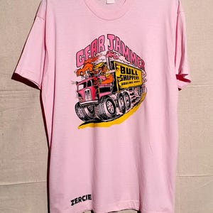 Può includere: T-shirt rosa chiaro con una grafica a fumetti di un camion e le scritte "GEAR JAMMER" e "BULL SHIPPERS HAULING ASSOC." La maglietta ha anche la scritta "ZERCIE" stampata in basso.