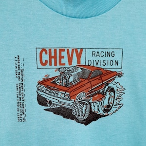 以下が含まれることがあります： 赤いシボレーのマッスルカーのグラフィックと「CHEVY RACING DIVISION」の文字が入った水色のTシャツ。シャツにはScreen Stars Bestのタグが付いており、アメリカ製です。