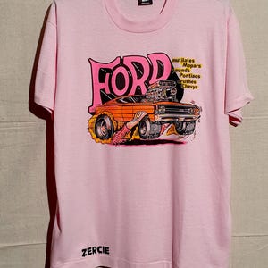 Può includere: T-shirt rosa chiaro con una grafica di un'auto muscolare Ford arancione vintage con fiamme e la scritta "FORD". La maglietta presenta anche il testo "mutilates, pounds, crushes, etc." e il nome "ZERCIE".