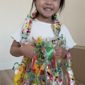 May include: A white short-sleeved ribbed shirt worn with a handmade lei or garland constructed from numerous small, individually wrapped snack bags in various colors, tied together with green ribbons and draped around the neck and torso.
