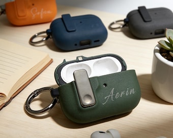 May include: Several coloured AirPod cases are displayed, including orange, navy, and green. The green case is open, showing the inside, and has the name "Aerin" written on it. Each case has a metal clasp and a small loop for attachment.