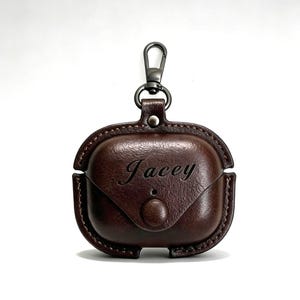 May include: A brown leather AirPod case with the name "Jacey" engraved on the front. The case has a snap closure and a metal clasp for attaching to a bag or keychain. The case is designed to protect and carry wireless earbuds.