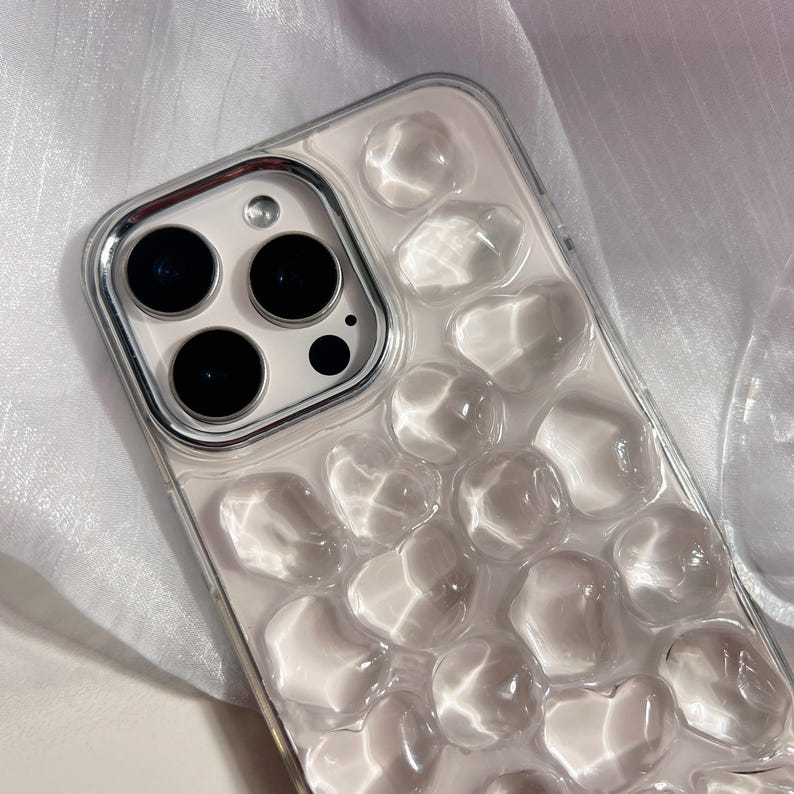 May include: Clear phone case with a bubbly, textured design. The case has a silver camera border and a light pink interior. The clear bubbles create a unique, eye-catching aesthetic. The case is designed to fit a smartphone.