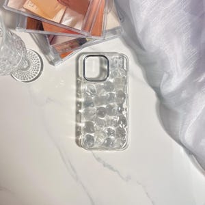 May include: Clear phone case with a textured, bubbly design. The case has a silver-colored camera ring and is set on a white marble surface. Other items in the background include a glass, and a stack of clear plastic cases.