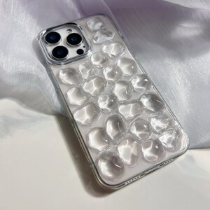 May include: Clear phone case with a pebble design. The case is transparent, allowing the phone's color to show through. The pebble design is raised and adds texture to the case. The camera lenses are visible.
