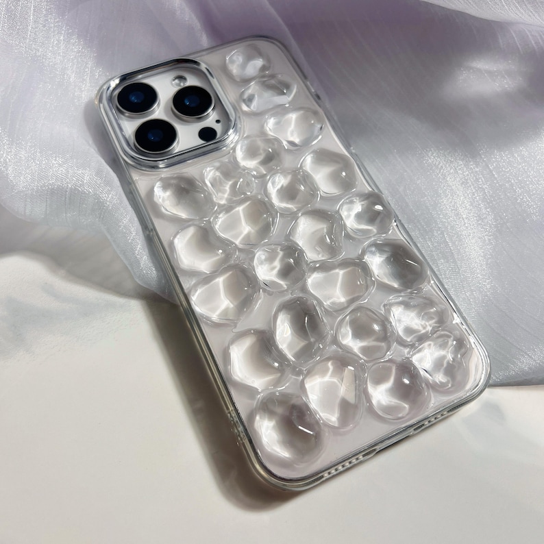 May include: Clear phone case with a pebble design. The case is transparent, allowing the phone's color to show through. The pebble design is raised and adds texture to the case. The camera lenses are visible.