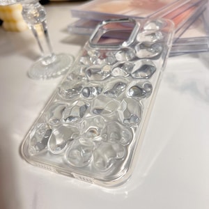 May include: Clear phone case with a textured, three-dimensional design. The case has a raised, irregular pattern of clear, rounded shapes. The phone case is made of a transparent material and has a silver-colored frame.