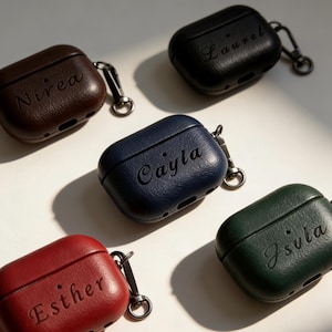 May include: A collection of personalized AirPod cases in various colors, including brown, black, navy blue, red, and green. Each case features a name engraved on the front and includes a silver-tone carabiner clip for attachment.