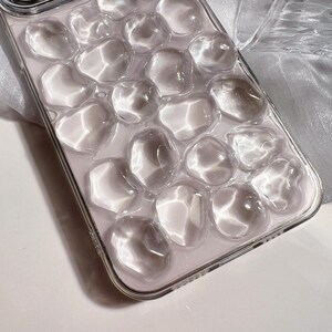 May include: Clear phone case with a bubbly, textured design. The case has a transparent appearance with rounded, irregular shapes, creating a unique, three-dimensional effect. The case is made of a clear material and has a shiny finish.