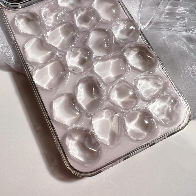 May include: Clear phone case with a bubbly, textured design. The case has a transparent appearance with rounded, irregular shapes, creating a unique, three-dimensional effect. The case is made of a clear material and has a shiny finish.