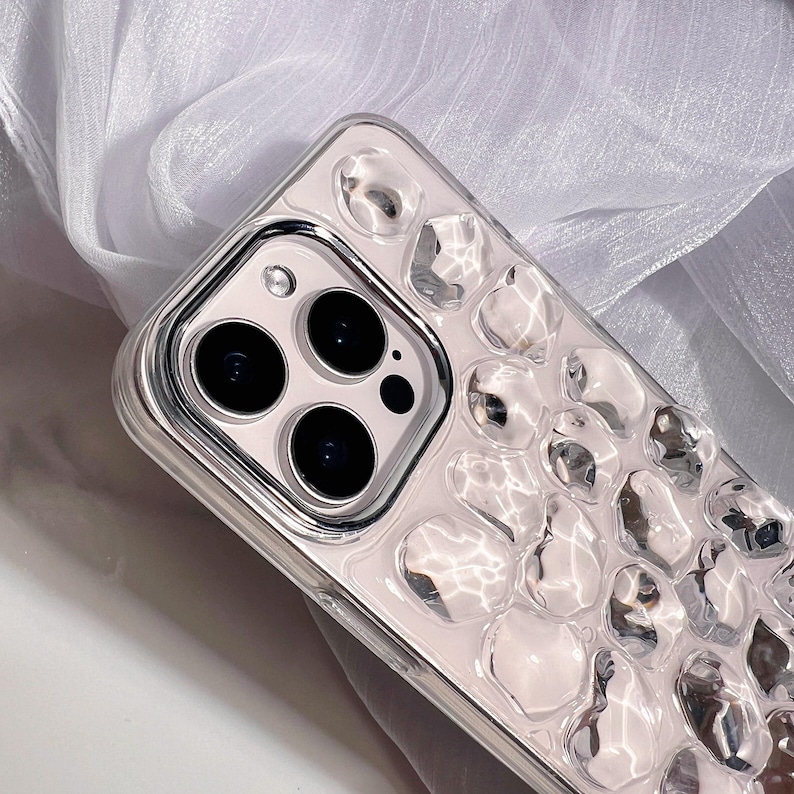 May include: A clear phone case with a silver frame and a textured surface of clear, rounded shapes. The case has a camera cutout with three black lenses and a flash. The background is a white, textured fabric.