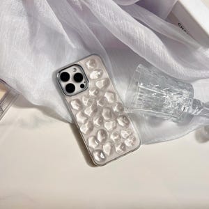 May include: A clear phone case with a textured, bubble-like design. The case is silver-edged and sits next to a clear, faceted glass. The background features a white, sheer fabric.