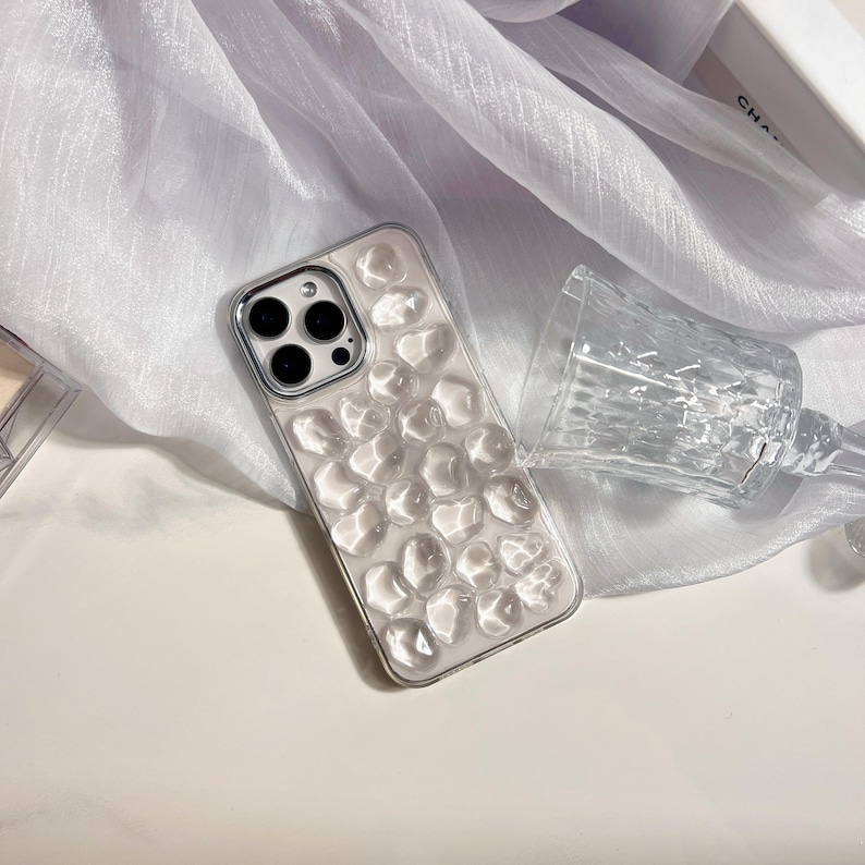 May include: A clear phone case with a textured, bubble-like design. The case is silver-edged and sits next to a clear, faceted glass. The background features a white, sheer fabric.