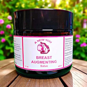 May include: A glass jar with a black lid, labeled "A New You Breast Augmenting Salve." The label features a pink graphic and text. The jar is 2 oz. and sits on a wooden surface with a blurred green background.