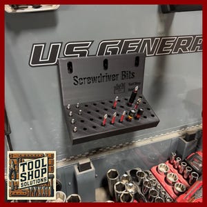 May include: A black screwdriver bit organizer with the text "Screwdriver Bits" and "Tool Shop Solutions". The organizer is mounted on a gray surface and holds various screwdriver bits with red and silver accents.