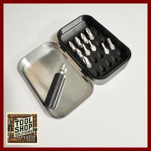 May include: A metal toolbox with a black interior and silver bits. The toolbox is open, revealing a set of silver screwdriver bits and a black handle. The toolbox is on a white surface. The toolbox has the text "TOOL SHOP SOLUTIONS".