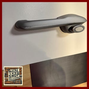 May include: A gray door handle with a rounded grip and a circular end. The handle is mounted on a light gray surface. The Tool Shop Solutions logo is in the bottom left corner.