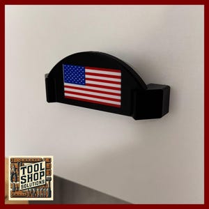 May include: A black wall-mounted tool holder with an American flag design. The holder features a semi-circular top and two rectangular side supports. The flag has red and white stripes with a blue field of stars.