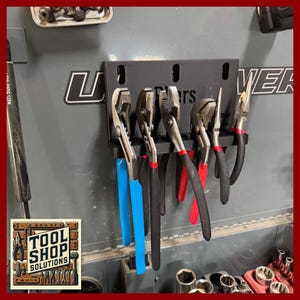 May include: A black metal tool holder with several pliers hanging from it. The pliers have blue, red, and black handles. The text "Pliers" is visible on the holder. The Tool Shop Solutions logo is in the lower left corner.