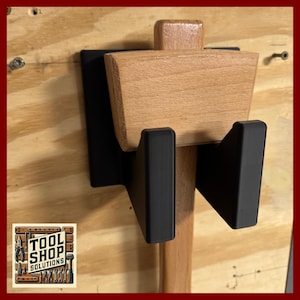 May include: A wooden mallet holder with black metal supports, mounted on a light brown wooden surface. The holder features a rectangular wooden head and a cylindrical handle. The Tool Shop Solutions logo is in the lower left corner.