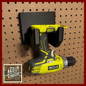 May include: A black drill holder mounted on a pegboard, holding a yellow and black Ryobi drill. The drill holder is made of plastic and has a rectangular backplate. The pegboard is brown with evenly spaced holes. The Tool Shop Solutions logo is in the lower left corner.