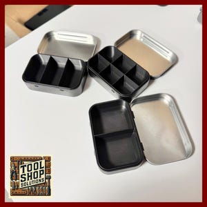 May include: Several small black plastic organizers with dividers sit open and closed next to silver metal tins. The organizers have different compartment configurations. The Tool Shop Solutions logo is in the lower left corner.