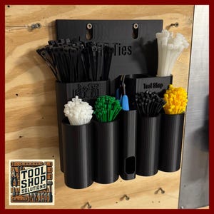 May include: Black plastic cable tie organizer mounted on a wooden surface. The organizer has multiple cylindrical compartments filled with white, green, black, and yellow cable ties. The word "Ties" is visible on the organizer. A logo for "Tool Shop Solutions" is in the lower left corner.