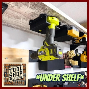 May include: Black under-shelf drill holder with a yellow and gray drill attached. The image shows a workshop setting with other tool holders and drills. The text "TOOL SHOP SOLUTIONS" is visible on a wooden sign. The text "*UNDER SHELF*" is also visible.