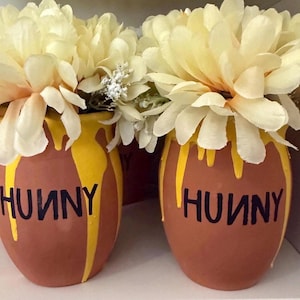 May include: Two terracotta-coloured pots featuring the word HUNNY in black lettering. Each pot has yellow paint dripping down from the rim and is filled with large, pale yellow artificial flowers and small white floral accents.