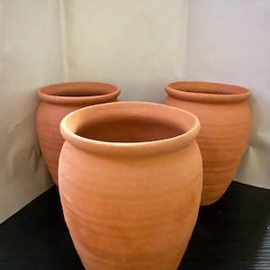 Könnte beinhalten: Drei unglasierte Keramiktöpfe in Terrakotta-Farbe. Die Töpfe haben einen abgerundeten Körper und eine breite, offene Mündung. Die Töpfe sind in einer Gruppe angeordnet, mit einem Topf im Vordergrund und zwei dahinter. Geeignet für den Innen- und Außenbereich.
