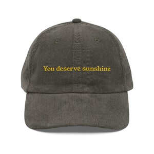 May include: A corduroy baseball cap in a muted olive green color. The cap features the embroidered text "You deserve sunshine" in a bright yellow color. The cap has a curved brim and a top button.