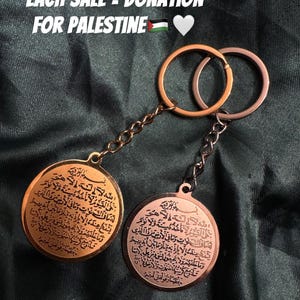 Könnte beinhalten: Zwei goldene und kupferne Schlüsselanhänger mit runden Metallanhängern, die mit arabischer Kalligraphie verziert sind. Der Text auf den Anhängern lautet "Allah la ilaha illa huwa lahu mulk" und "Allah la ilaha illa huwa lahu mulk". Die Schlüsselanhänger befinden sich auf einem dunkelgrünen Stoffhintergrund. Der Text "EACH SALE = DONATION FOR PALESTINE" befindet sich oben im Bild.