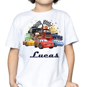 May include: White t-shirt with a vibrant graphic of animated race cars, with a red car prominent at the front. The name "Lucas" is printed in navy blue script below the cars. The design includes checkered flags and stars.
