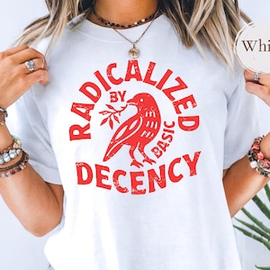 May include: White t-shirt with a red graphic that reads "RADICALIZED BY BASIC DECENCY." The graphic features a red bird holding a sprig. The shirt is a crew neck.