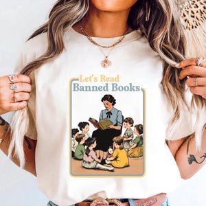 May include: White t-shirt with a vintage-style illustration of a teacher reading to a group of children. The text "Let's Read Banned Books" is printed in yellow and blue on the shirt.
