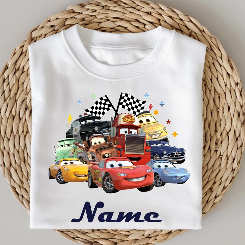May include: White t-shirt with a vibrant graphic of cartoon cars, including Lightning McQueen. The design incorporates checkered flags and the word "Name" in navy blue script. The cars are arranged in a playful, dynamic composition.