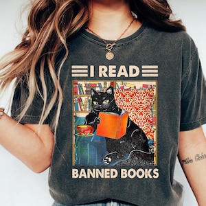 May include: A dark gray t-shirt with a black cat reading a book in a bookshelf setting. The text on the shirt reads "I read banned books".