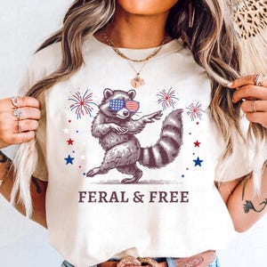 May include: Cream-colored t-shirt featuring a raccoon wearing American flag sunglasses, dancing with fireworks and stars. The text "FERAL & FREE" is printed below the raccoon. The design is in red, white, and blue.