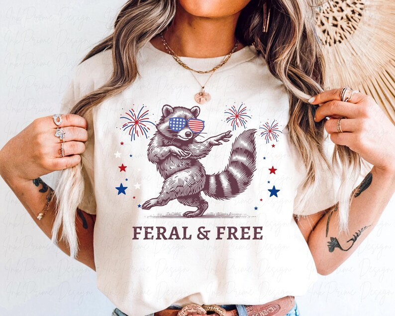 May include: Cream-colored t-shirt featuring a raccoon wearing American flag sunglasses, dancing with fireworks and stars. The text "FERAL & FREE" is printed below the raccoon. The design is in red, white, and blue.