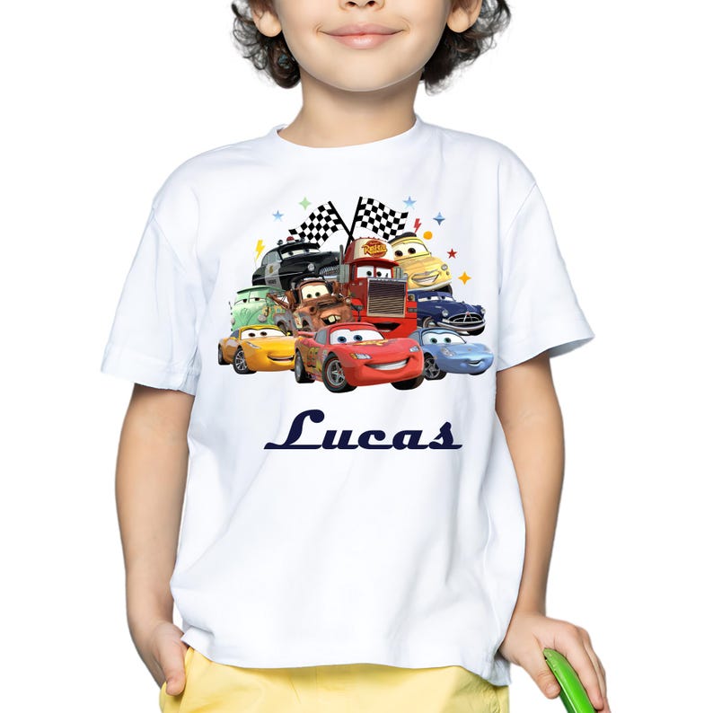 May include: White t-shirt with a vibrant graphic of animated race cars, with a red car prominent at the front. The name "Lucas" is printed in navy blue script below the cars. The design includes checkered flags and stars.
