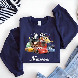 May include: Navy blue sweatshirt with a vibrant graphic of cartoon cars, including Lightning McQueen. The design incorporates checkered flags and stars. The word "Name" is written in white script below the cars. The sweatshirt is made of a soft material.